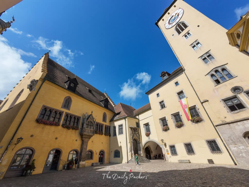 Regensburg Old Town Hall with its yellow facade, clock tower, and medieval courtyard under a bright blue sky.