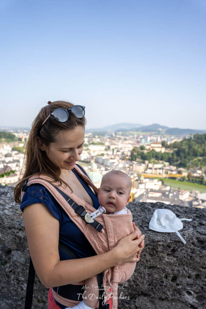 Femme tenant un bébé dans un porte-bébé au point de vue de la forteresse de Hohensalzburg avec la ville de Salzbourg en arrière-plan.