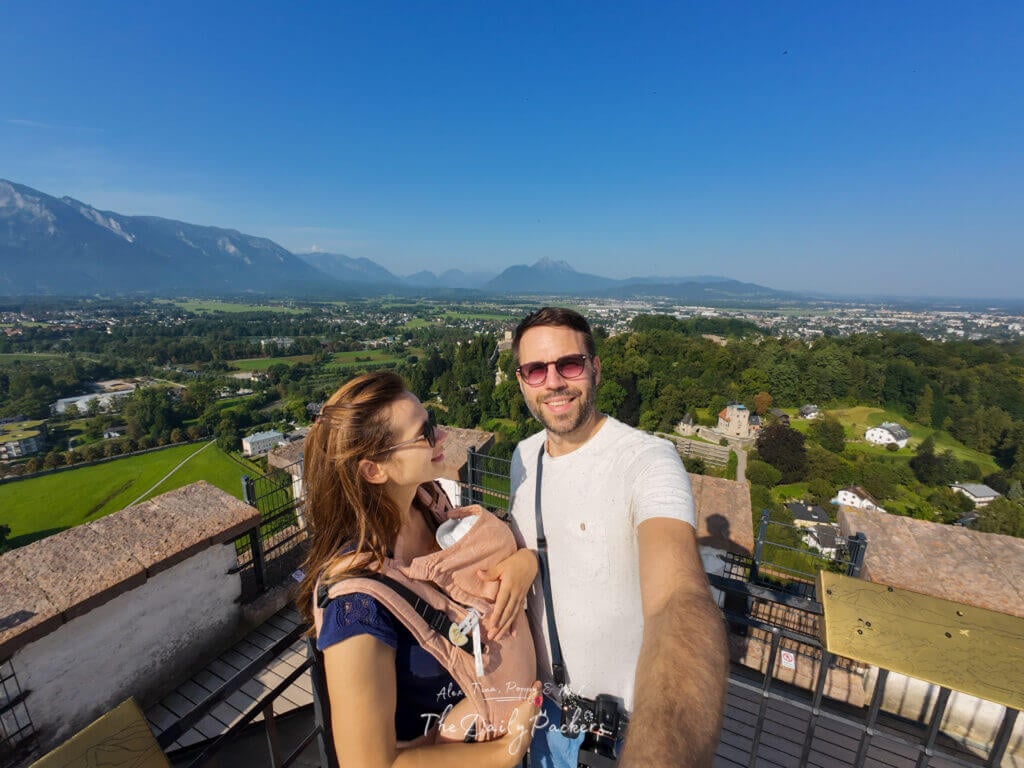 Couple with baby taking a selfie from a tower viewpoint at Hohensalzburg Fortress overlooking Salzburg and surrounding mountains.