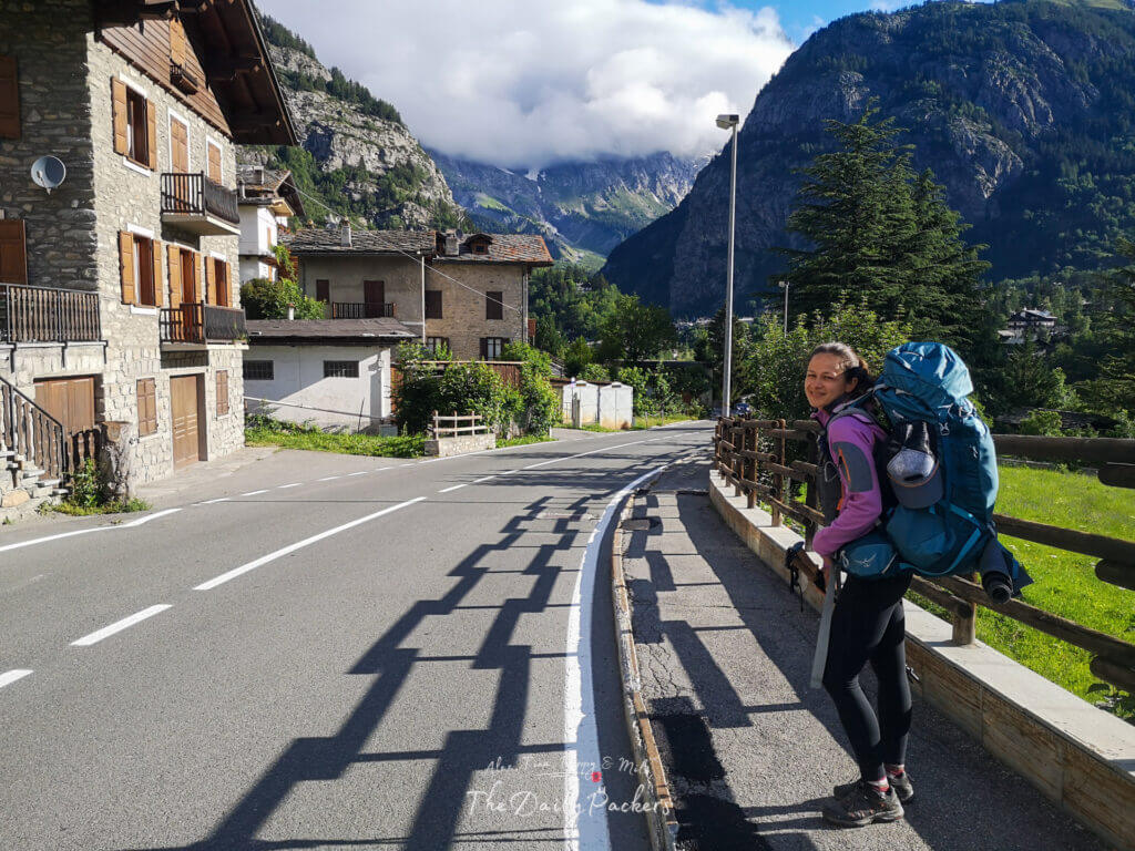 Woman with a large backpack walking through a village street in Chamonix with stone houses, green mountains, and clouds in the background.