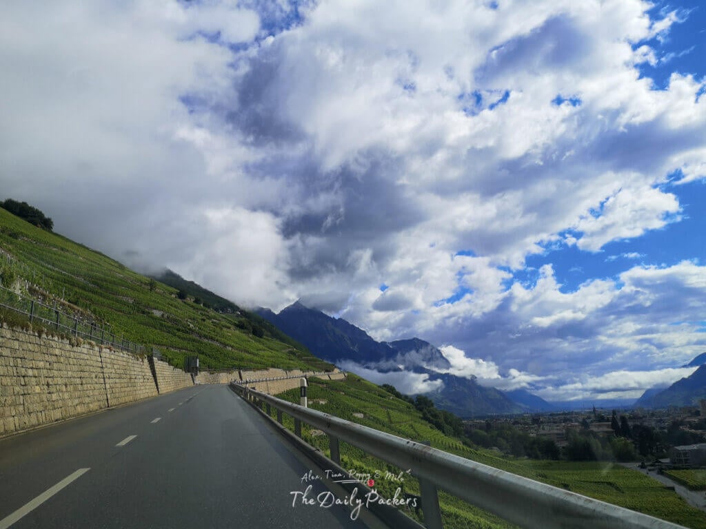 Open road winding through vineyard slopes with a view of the valley and mountains partly covered in clouds.