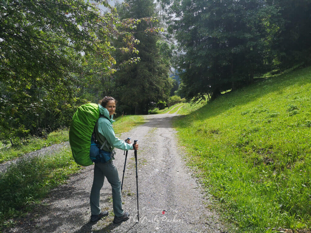 Female hiker with a green rain cover on her backpack walking up a gravel path through green meadows.