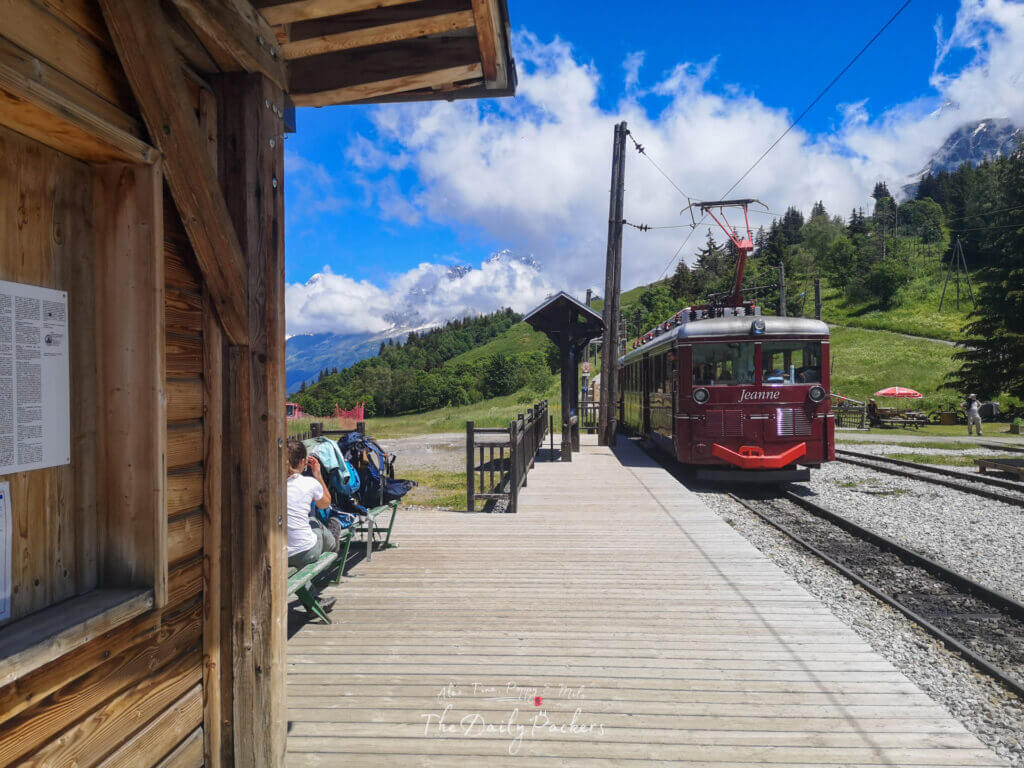 Mountain train station at Col de Voza with hikers resting and the red Jeanne train under a bright sky.