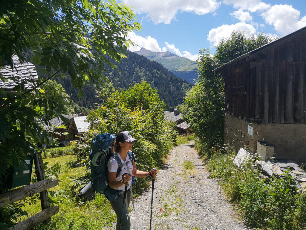 Hiker walking along a quiet alpine path lined with chalets and greenery, carrying a large backpack.