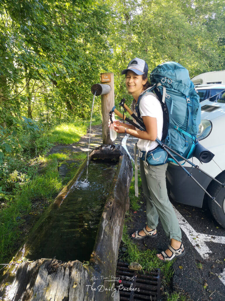 Randonneur souriant remplissant de l'eau à une fontaine en bois le long du sentier près des Contamines.