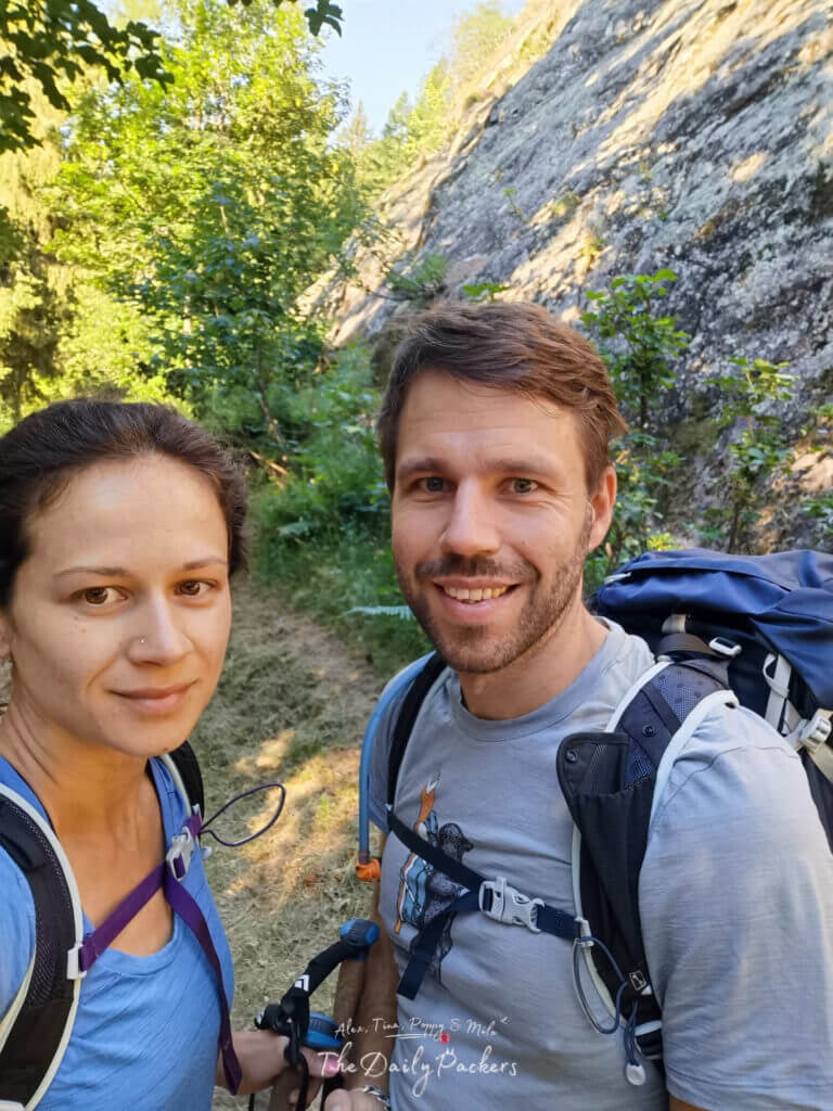 Trail selfie of two hikers beneath a granite wall and trees
