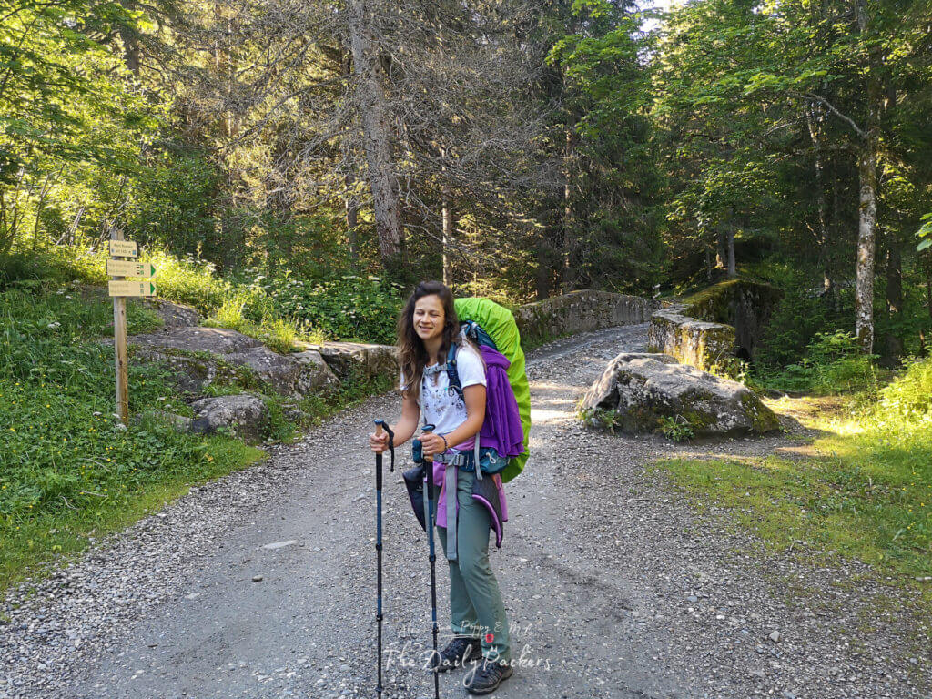 Randonneur avec un grand sac à dos traversant un sentier forestier près d'un pont en pierre sur le chemin du Refuge de la Balme.