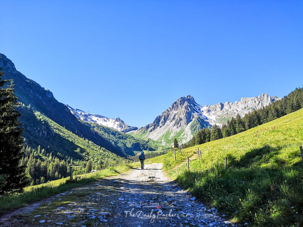 Hiker walking along a wide mountain trail with stunning peaks and clear blue sky on the way to Refuge Nant Borrant during the TMB Stage 2 from Les Contamines to Col de la Croix du Bonhomme