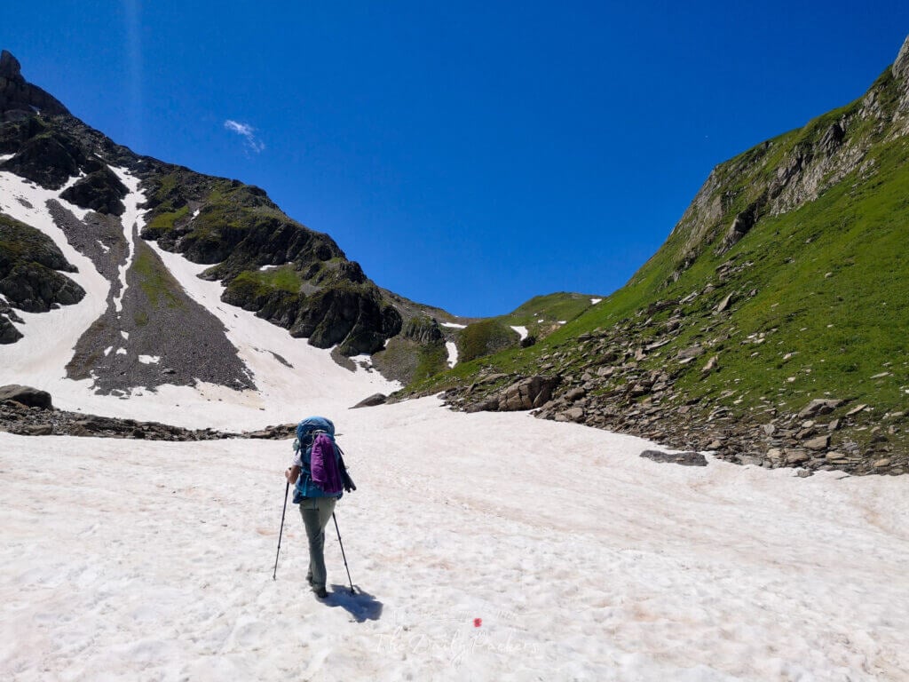 Col de montagne enneigé avec des randonneurs traversant et un petit abri en bois sur la crête.