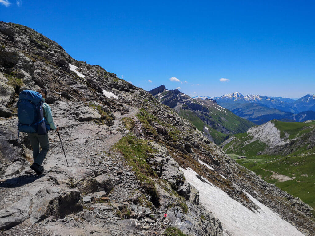 Randonneur marchant le long d'un sentier de montagne rocailleux avec des plaques de neige et des vues panoramiques sur les Alpes.