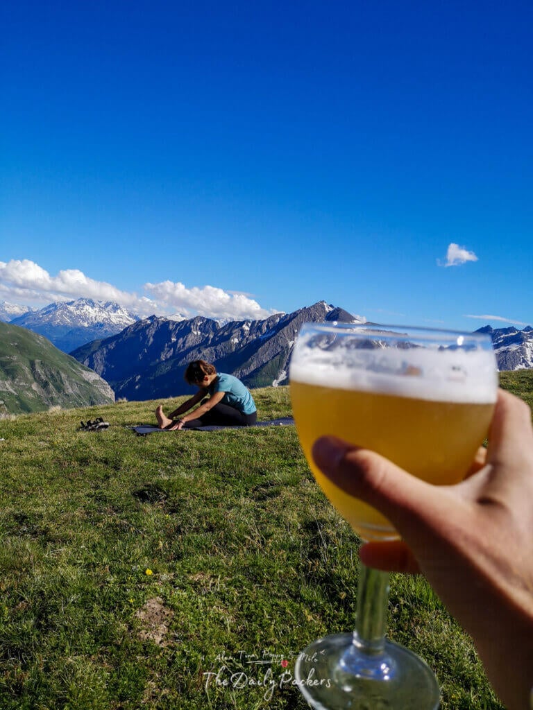 Main tenant un verre de bière avec des montagnes en arrière-plan, tandis qu'une personne s'étire sur un tapis de yoga dans l'herbe.