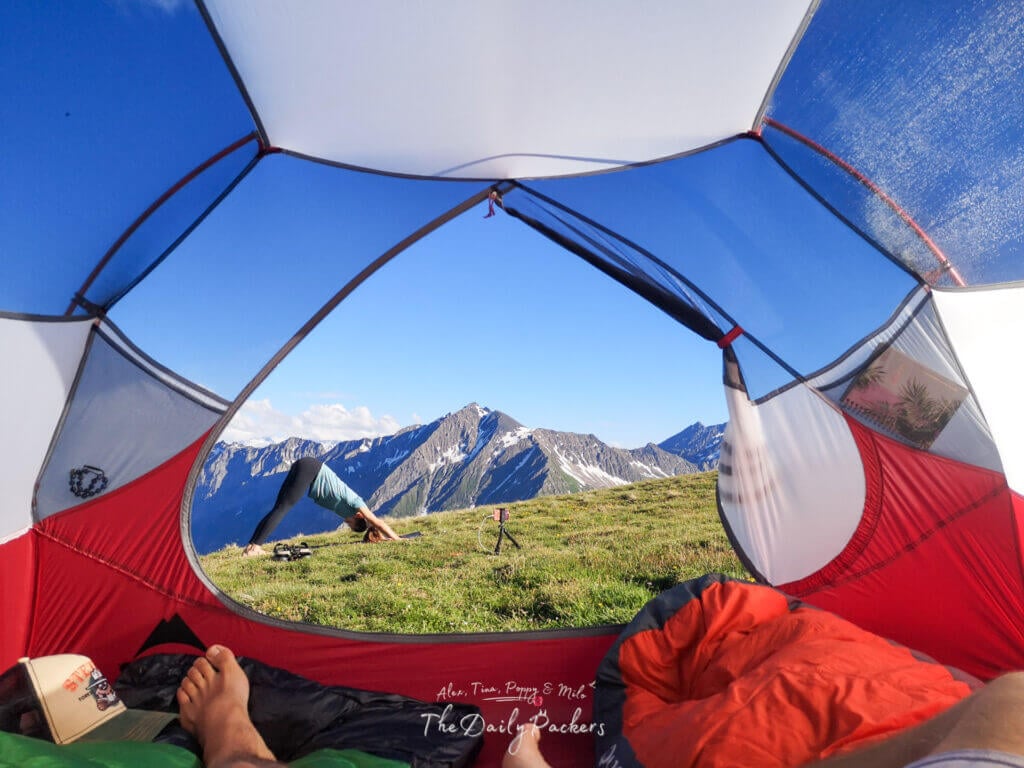 View from inside a tent looking out to the mountains, with a person practicing yoga outdoors under a clear blue sky.