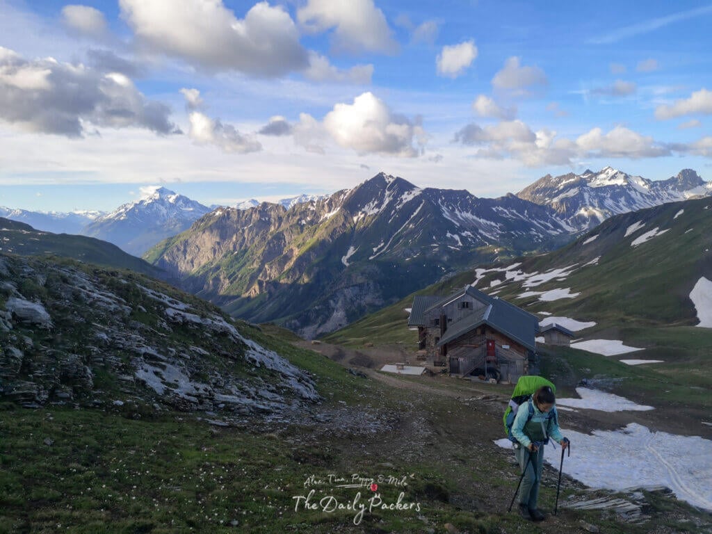 Randonneur quittant un refuge de montagne sur une pente herbeuse avec des pics dramatiques et des plaques de neige.