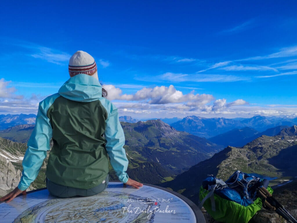 Randonneur assis sur une table d'orientation au sommet, regardant les vallées et les chaînes de montagnes sous un ciel bleu éclatant.