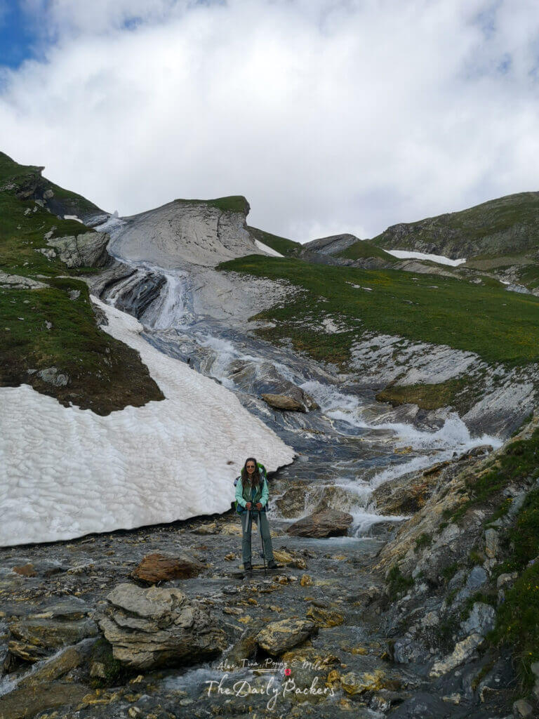 Randonneur traversant un ruisseau rocheux avec des plaques de neige et une cascade coulant le long de la pente de la montagne.