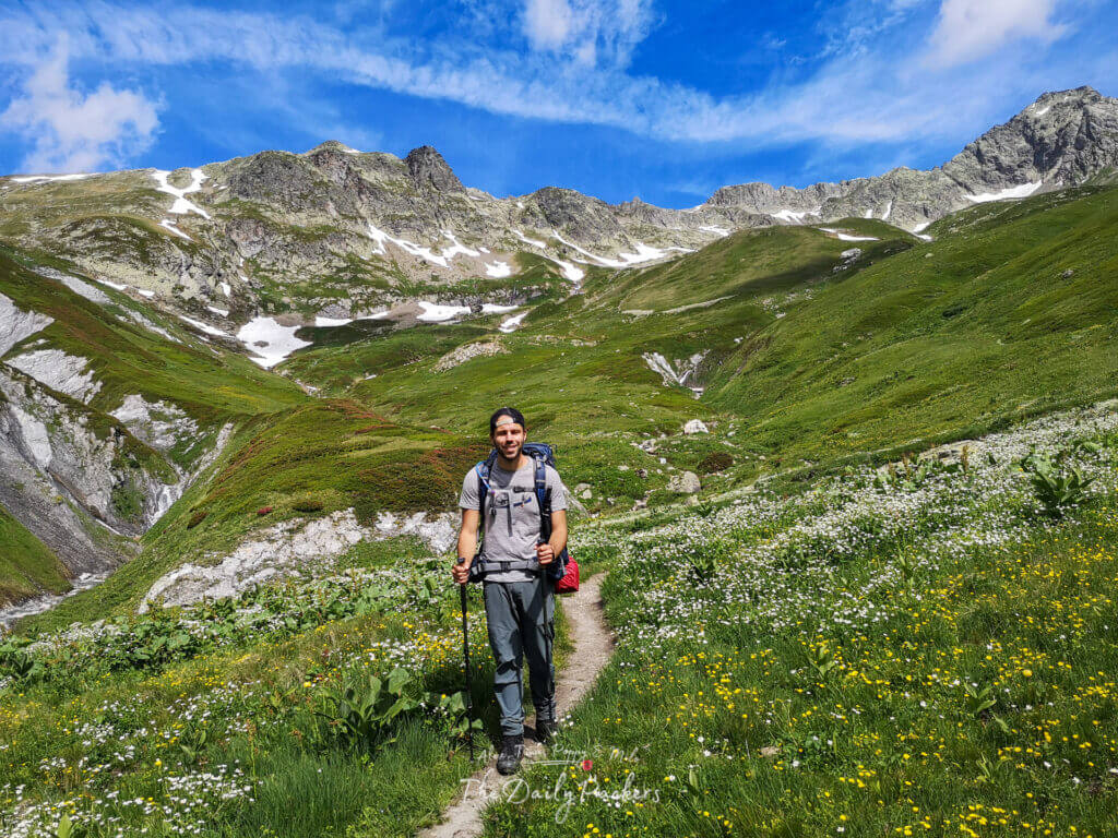 Randonneur debout sur un sentier alpin entouré de fleurs sauvages, avec des pentes vertes abruptes et des pics rocheux derrière.