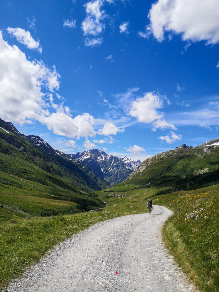Randonneur marchant sur une large route de gravier serpentant à travers une vallée avec des sommets enneigés devant.