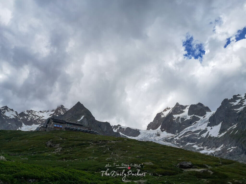 Refuge de montagne situé sur une crête avec des sommets enneigés et des glaciers en arrière-plan.