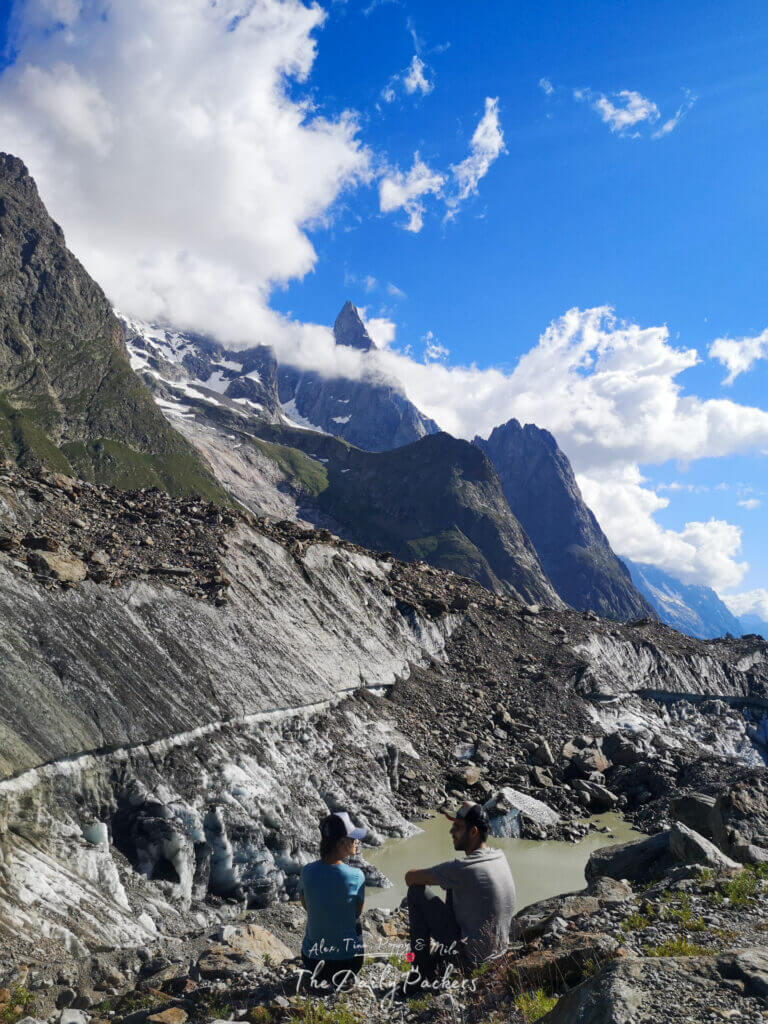 Two hikers sitting on rocks near a glacier lake with jagged peaks and clouds in the background.