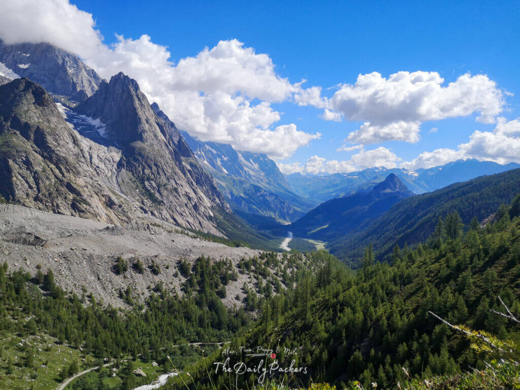 Vue large sur une vallée alpine avec des pics rocheux dentelés, des forêts vertes et une rivière sinueuse en contrebas.