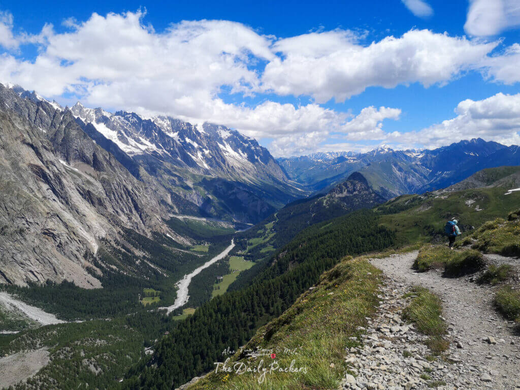 Backpacker hiking on a rocky path overlooking a vast valley with snow-capped mountains.