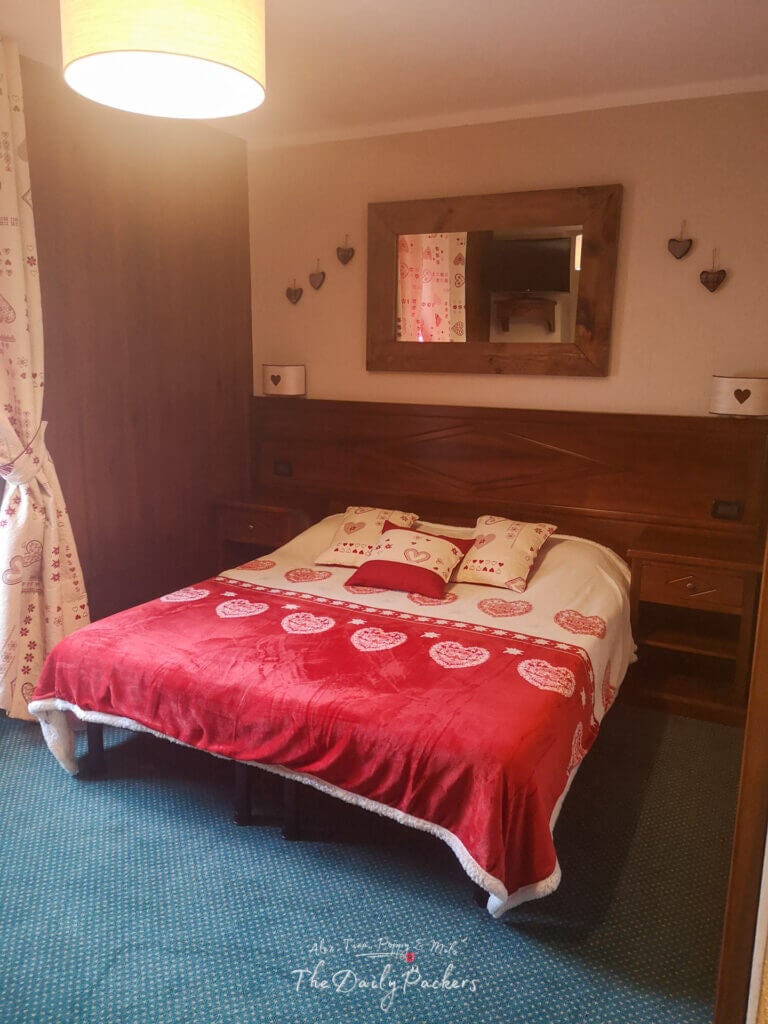 Chambre d'hôtel confortable avec un couvre-lit rouge décoré de cœurs, des meubles en bois et un éclairage chaleureux.