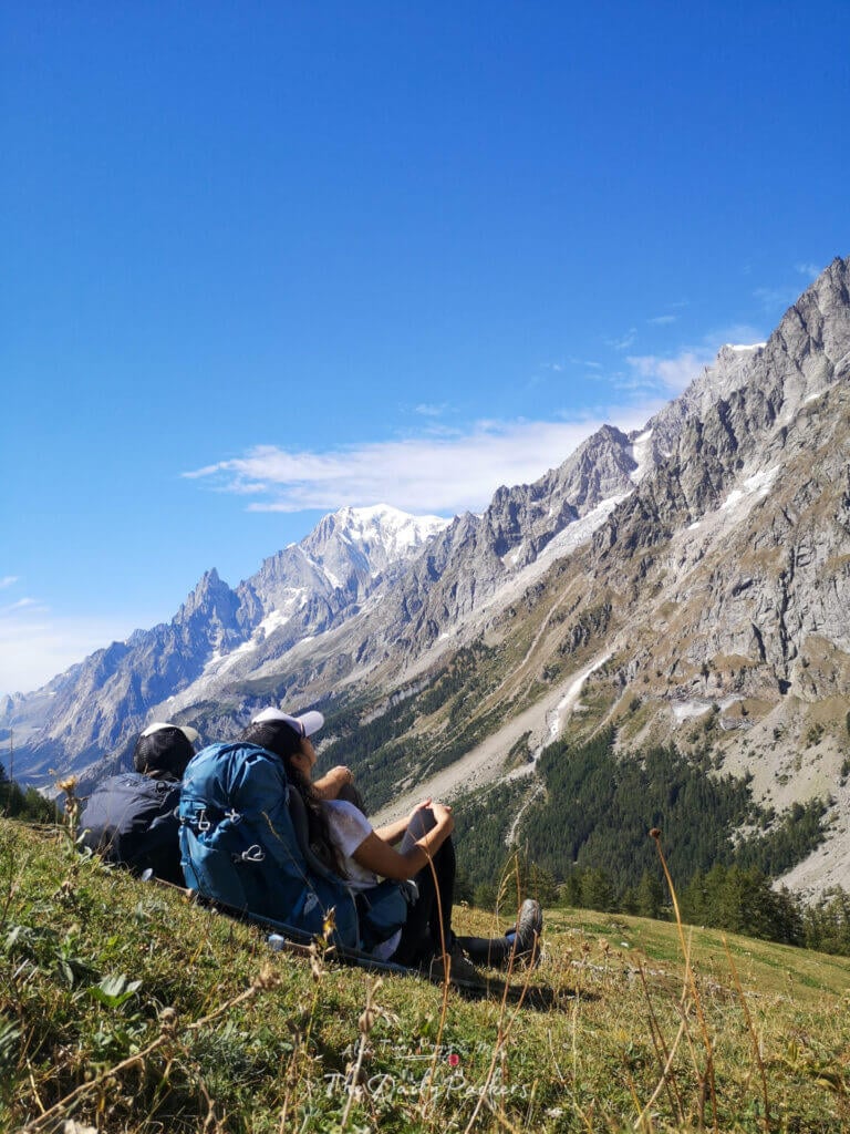 Couple resting on the grass with backpacks, enjoying panoramic views of Mont Blanc and surrounding peaks.