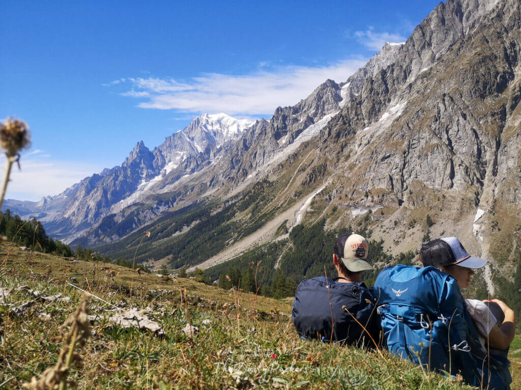Two hikers sitting with backpacks on the grass, overlooking the Mont Blanc massif under a clear blue sky during the TMB Stage 5 from Courmayeur to Rifugio Bonatti