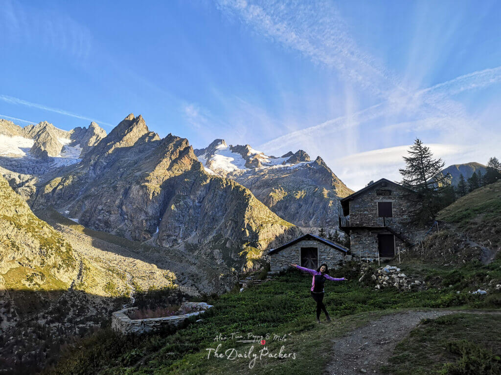 Randonneur posant joyeusement à côté de vieilles maisons alpines en pierre avec des glaciers en arrière-plan sur l'étape 6 du TMB du refuge Bonatti à La Fouly