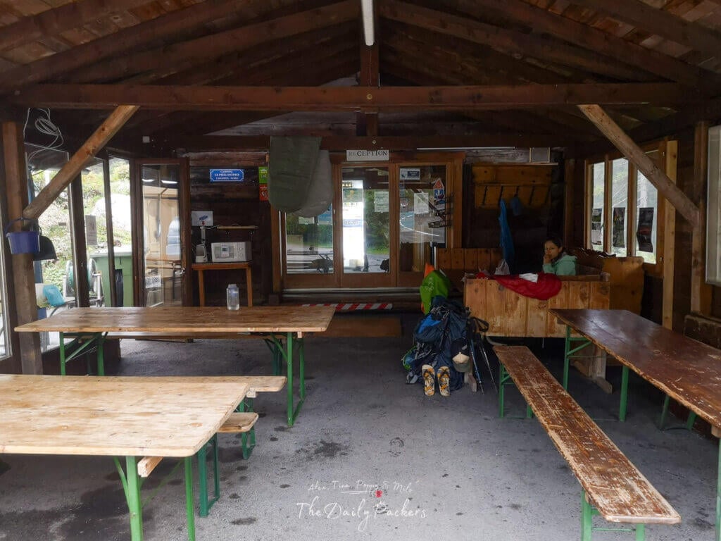 Wooden shelter with picnic benches and hikers resting inside at a campground reception area.