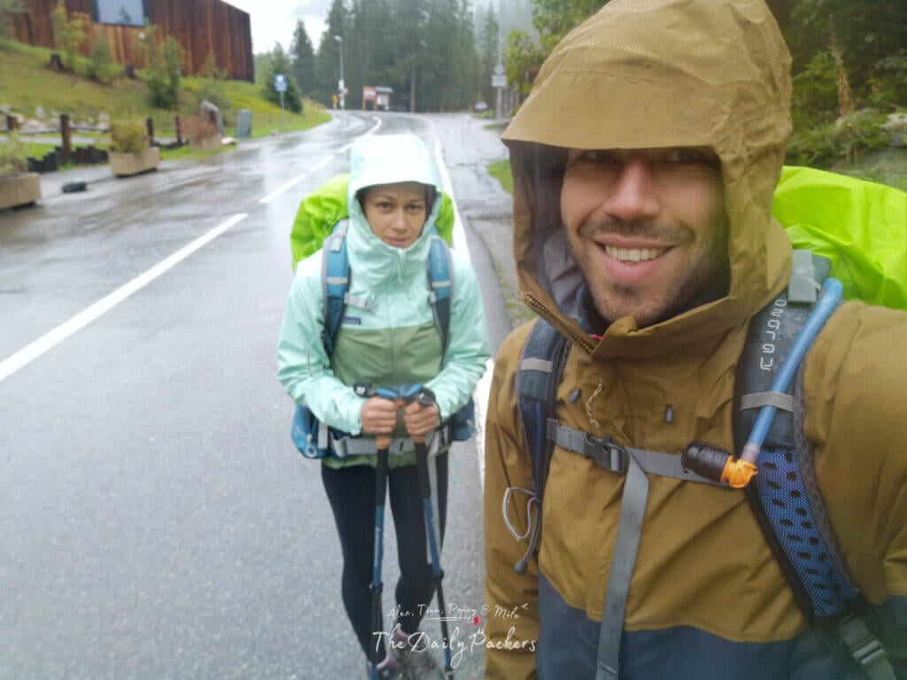 Two hikers in rain jackets with backpacks smiling at the camera on a wet road in the mountains.