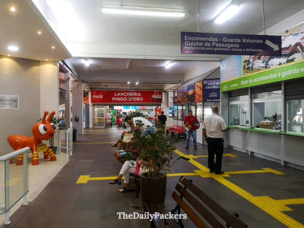Ticket counters and waiting area inside Pelotas bus terminal, with people seated and signage in Portuguese.