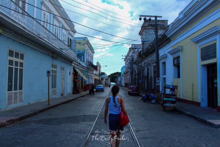 Woman walking along a cobblestone street in Cienfuegos, surrounded by pastel colonial buildings and vintage vehicles.