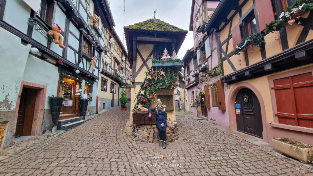 Peaceful corner in Eguisheim with festive decorations, teddy bears on windows, and cobblestone streets curving between pastel houses.
