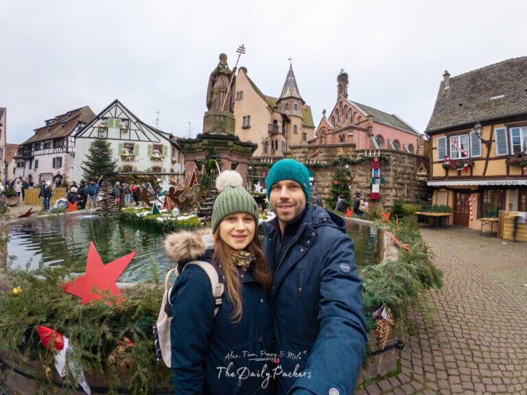 Couple taking a selfie at Saint-Léon Square during the Eguisheim Christmas Market, surrounded by holiday decorations, a fountain, and historic buildings