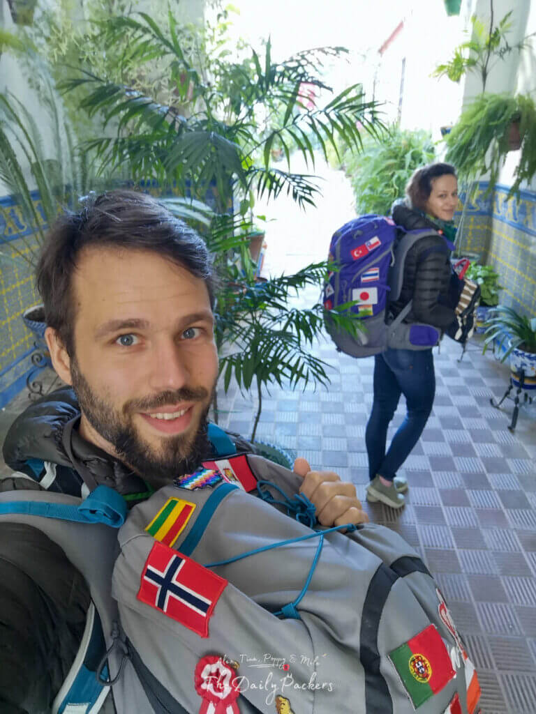 Voyageurs avec sacs à dos souriants dans une charmante cour andalouse décorée de plantes vertes et de carreaux traditionnels.