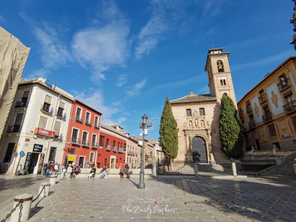 Plaza Nueva in Granada with colorful buildings, people relaxing in the square, and the Iglesia de San Gil y Santa Ana in the background.