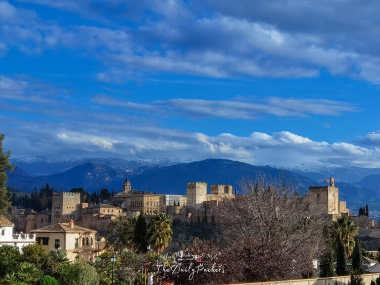 View of the Alhambra from a viewpoint in Granada with the Sierra Nevada mountains in the background under a bright blue sky.