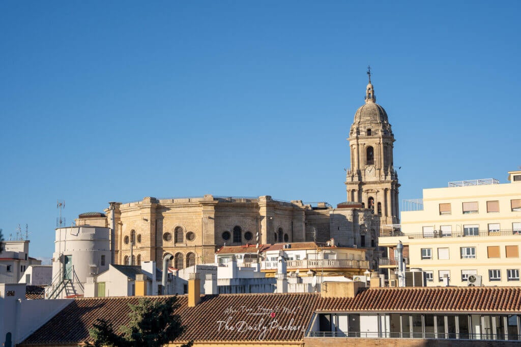 Scenic view of Málaga Cathedral rising above city rooftops, seen from the Alcazaba fortress on a sunny day.