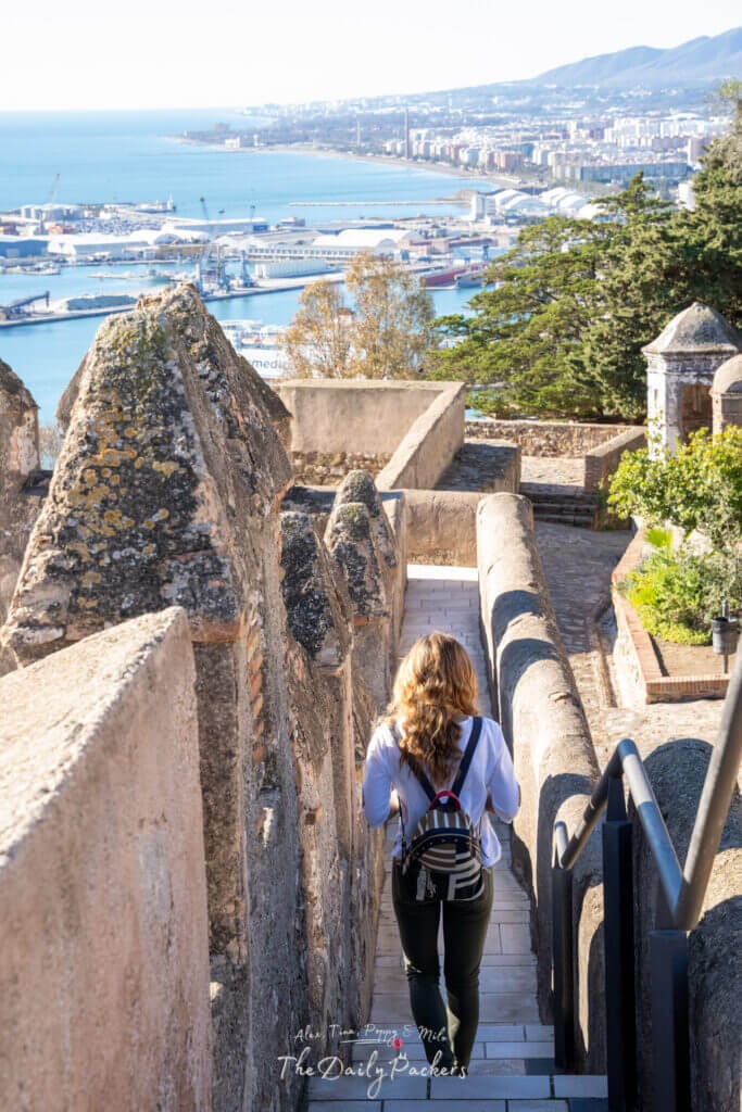 Traveler walking down the ancient walls of Gibralfaro Castle with panoramic views over Málaga’s port and coastline.