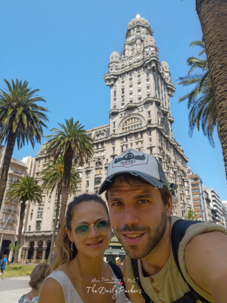 Couple taking a selfie in front of the Salvo Palace at Plaza Independencia under a bright blue sky in Montevideo.