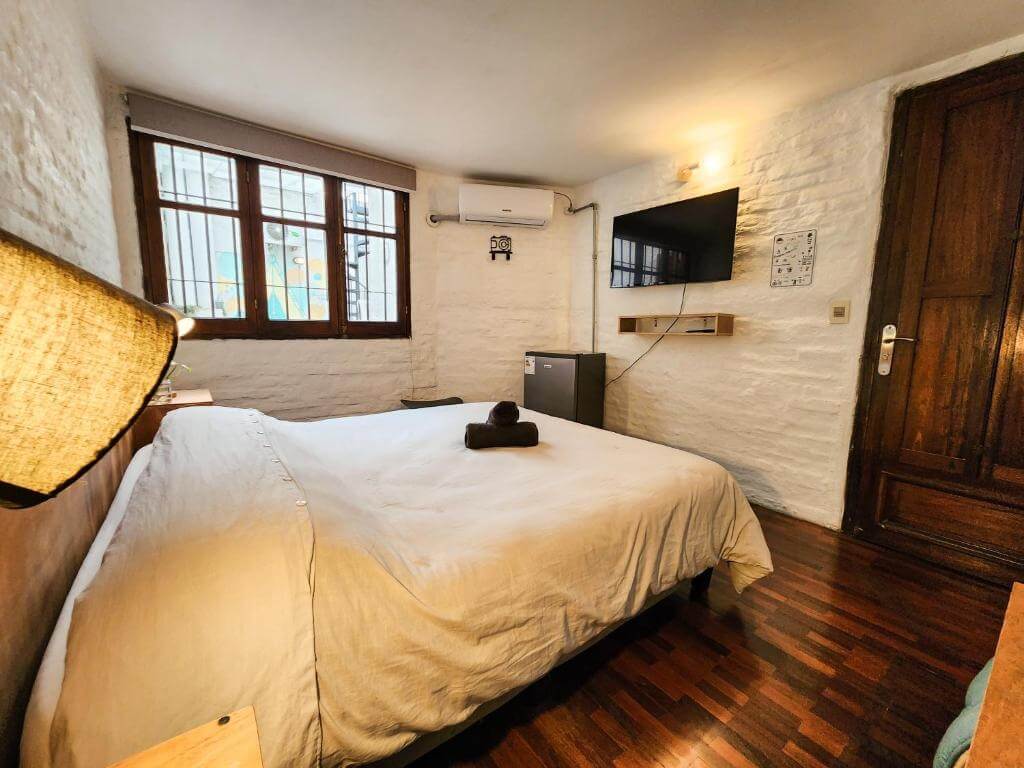 Bright double room at MedioMundo Hostel in Montevideo with rustic white brick walls, wooden floors, and a simple cozy setup.