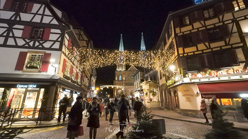 Night view of Rue du Chanoine Gyss in Obernai, glowing with hanging Christmas lights and the church towers in the background.