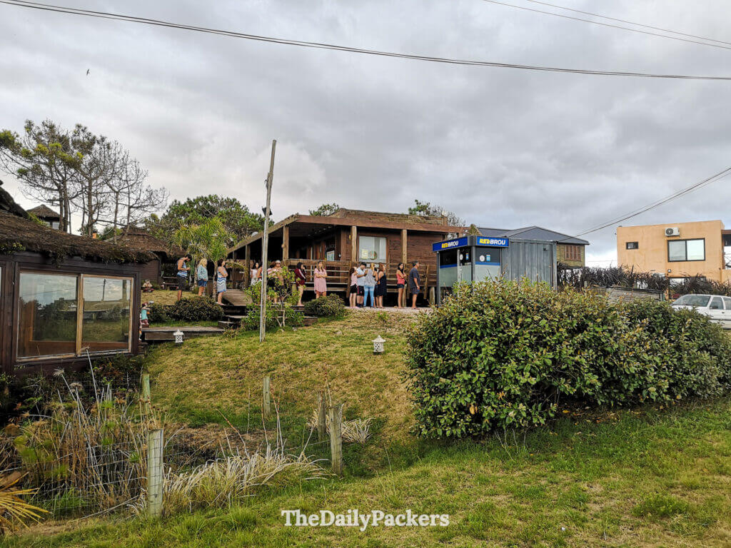 People queuing at an outdoor ATM next to rustic wooden buildings and cafes in the center of Punta del Diablo.