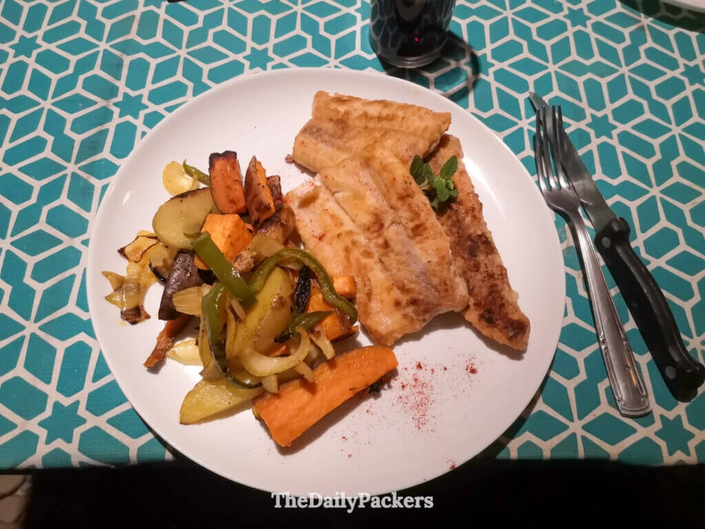 Gros plan d'une assiette avec du poisson grillé, des légumes rôtis et des patates douces servis sur une nappe turquoise à motifs.