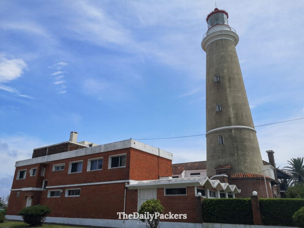 The iconic Punta del Este lighthouse standing tall beside red-brick buildings and palm trees under a clear sky.