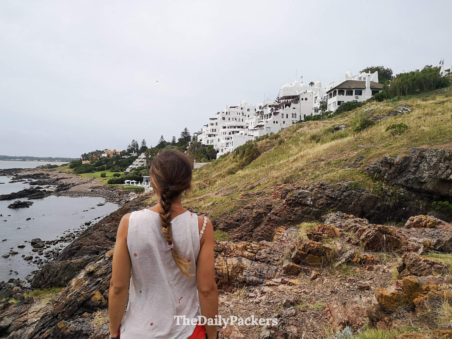 Woman standing on rocky cliffs gazing toward Casa Pueblo, surrounded by coastal scenery in Punta Ballena.