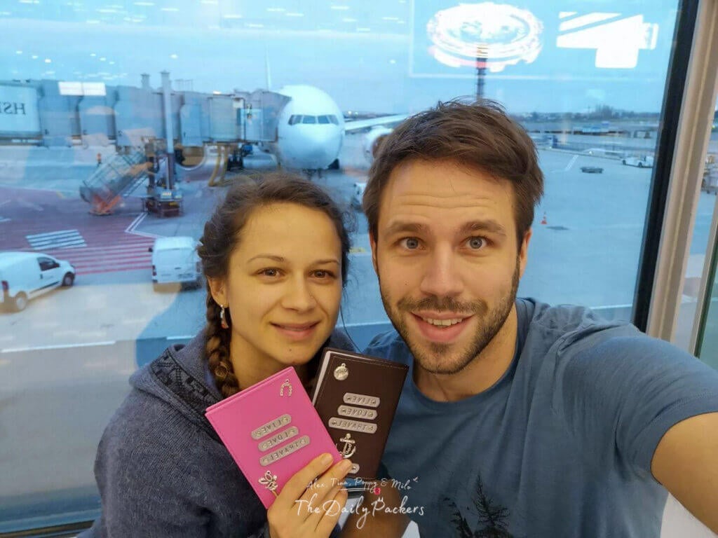 Couple smiling and holding their passports with an airplane and boarding gate in the background.