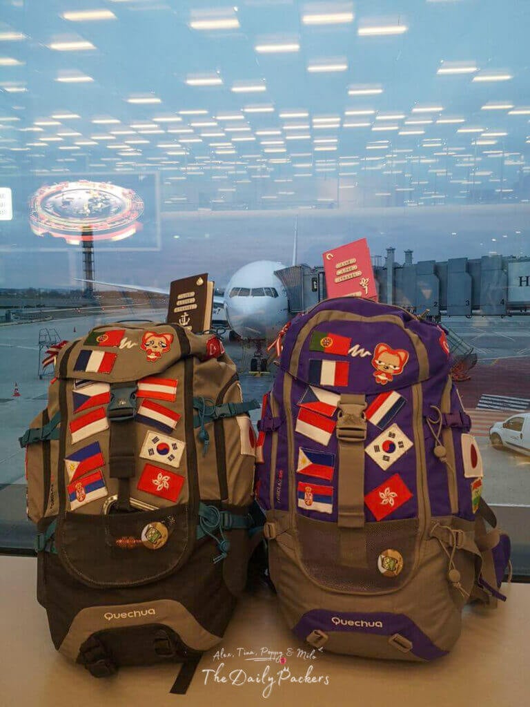 Two colorful travel backpacks decorated with country flag patches and passports placed on top, waiting at an airport gate.