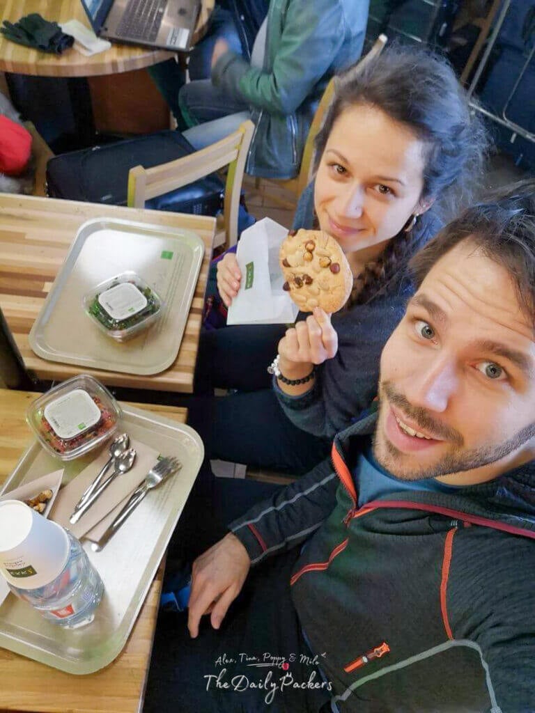 Couple sitting at a café with trays of food and drinks, smiling and enjoying a quick airport meal.